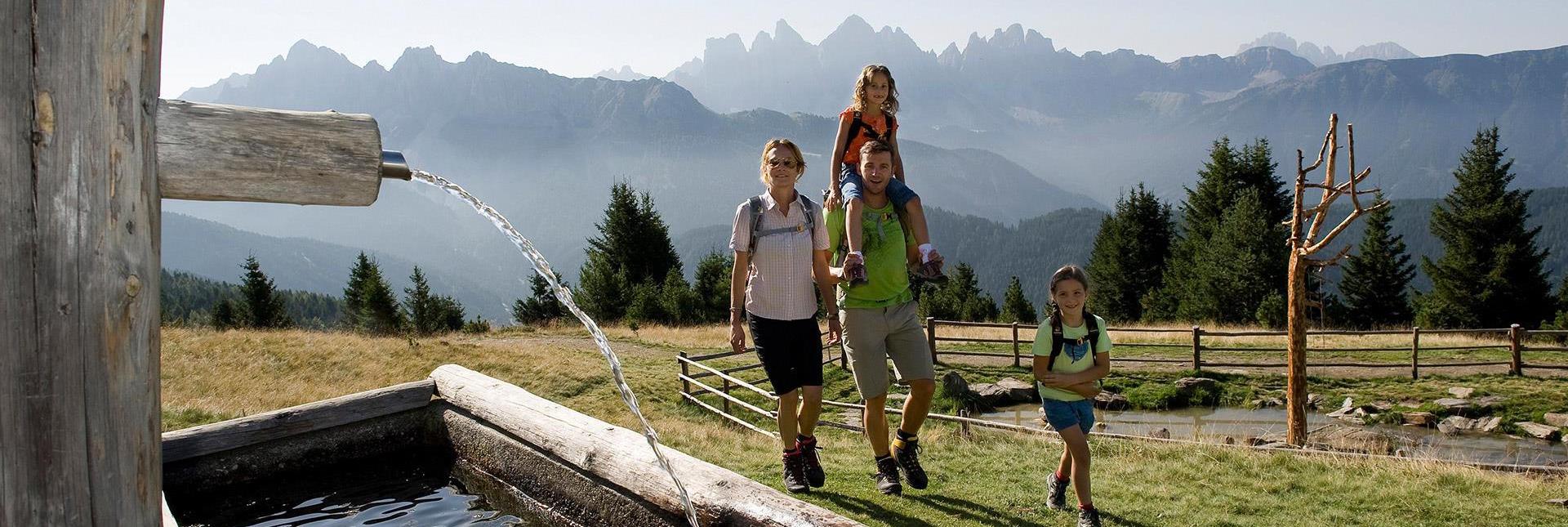 Una famiglia fa un'escursione accanto a una fontana nelle Dolomiti, con le imponenti vette delle Odle sullo sfondo.
