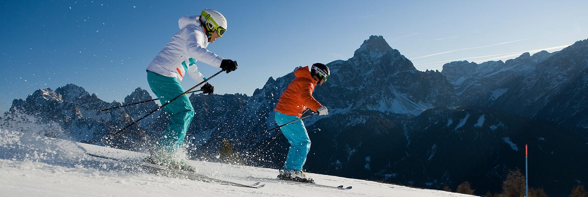 Due sciatori scendono una pista tra montagne innevate e cielo azzurro.